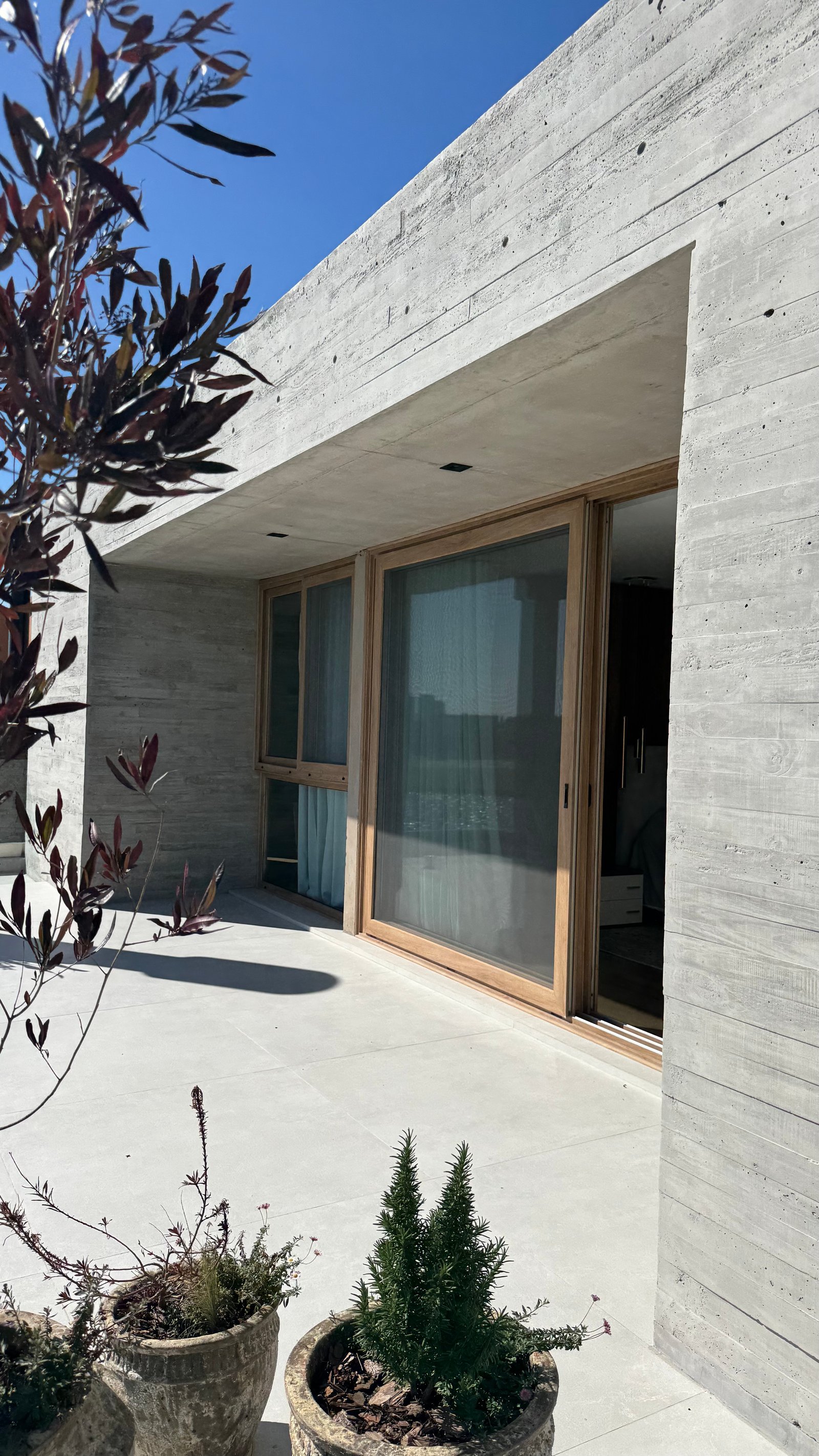 Fachada moderna de hormigón crudo con puertas de vidrio y patio de baldosas — proyecto CASA MHN, por Hermann Arquitectos, en Nordelta